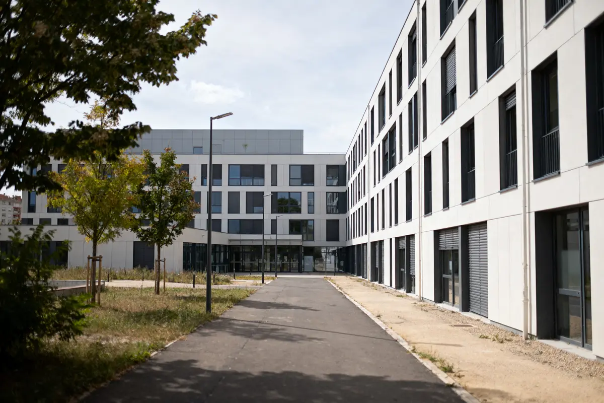Bâtiment moderne avec façade blanche et fenêtres, entouré d'arbres et d'un chemin pavé, par temps ensoleillé.