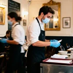Serveurs masqués et gantés travaillent dans un restaurant élégant, intérieur classique avec tables dressées.