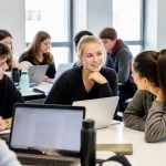 Groupe d'étudiants discutant autour d'une table, ordinateurs portables et bouteilles d'eau posés devant eux.