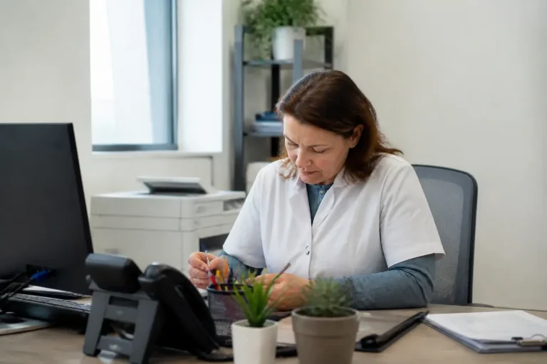 Femme en blouse blanche travaillant à un bureau avec ordinateur, téléphone et plantes vertes.